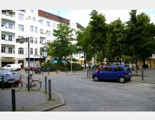 Straßenszene mit Kopfsteinpflaster in einem Wohnviertel, umgeben von weißen Mehrfamilienhäusern mit Balkonen. Im Vordergrund Fahrräder, geparkte Autos und eine kleine Grünfläche mit Bäumen; eine Uhr und Straßenschilder sind gut sichtbar.
