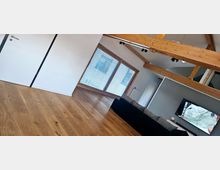 Wohnzimmer mit modernem Design, Holzdielenboden und sichtbaren Holzdeckenbalken. Eine große Fensterfront mit Lamellenblickschutz dominiert den Raum, daneben befinden sich eine weiße Tür, ein graues Sofa und ein an der Wand montierter Fernseher.