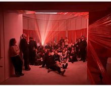 Ein Raum mit roten Lichtprojektionen und Wänden, die mit roter Plastikfolie bedeckt sind. Zahlreiche Personen sitzen oder stehen, teils an die Wand gelehnt, in einer konzentrierten Atmosphäre, während Lichtstrahlen von der Mitte des Raumes ausgehen.