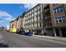 Straßenansicht einer städtischen Wohnhauszeile mit mehrstöckigen Altbaufassaden, darunter ein helles Gebäude mit reich verzierten Fensterrahmen, neben moderneren Häusern mit Balkonen. Entlang der breiten Fahrbahn stehen geparkte Autos und ein Lkw; am Erdgeschoss sind Schaufenster und ein kleiner Außenbereich erkennbar, darüber blauer Himmel mit Wolken.