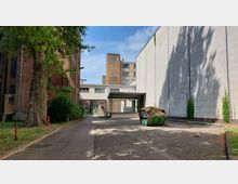 Industriegelände mit Backstein- und Betongebäuden, einem großen Baum links und einer Containermulde mit Gras oder Erde rechts. Im Hintergrund führt ein überdachter Übergang zwischen den Gebäuden.