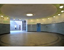 Runder, unterirdischer Durchgang mit blau gefliesten Wänden, Graffiti und einem blauen Türrahmen; links führt eine Treppe ins Freie, und rechts ist ein verziertes Metalltor zu sehen. Die Decke ist mit integrierten Lampen beleuchtet, und der Boden ist mit Steinfliesen bedeckt.