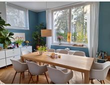 Esszimmer mit einer hellen Holztischgruppe, umgeben von weißen Stühlen, vor einer blauen Wand. Große Fenster lassen viel Tageslicht herein, mit Blick auf grüne Bäume; Dekorationen wie Pflanzen, Vasen und ein Sideboard mit Gegenständen runden den Raum ab.