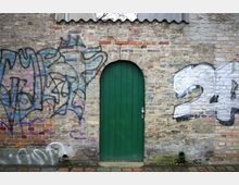 Backsteinwand mit grüner, schmaler Tür in Rundbogenform, flankiert von Graffiti auf beiden Seiten; im unteren Bereich gepflasterter Boden.