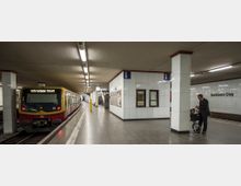 Eine U-Bahn-Station mit einem wartenden Zug der Linie S25 Richtung "Teltow Stadt" auf der linken Seite. Die Station ist modern gestaltet mit hellen Fliesenwänden, Beleuchtung von der Decke und einem Informationshäuschen in der Mitte; an der rechten Wand steht "Potsdamer Platz".