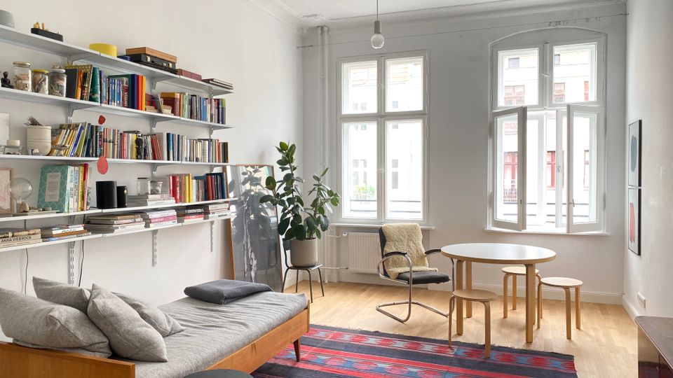 Helles Wohnzimmer mit hohen Decken, Stuck und zwei großen Sprossenfenstern, eines davon geöffnet. Links befinden sich wandmontierte Regale voller Bücher über einem Daybed; im Raum stehen ein runder Tisch mit Hockern, ein Schaukelstuhl, eine Zimmerpflanze und ein gemusterter Teppich auf Holzboden.