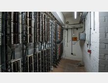 Ein schmaler technischer Raum mit blanken weißen Wänden und mehreren Reihen von metallischen Schaltkästen mit sichtbaren Kabeln. An der Wand befinden sich Verteilerkästen und Leitungen, der Boden ist aus graubraunem Beton.