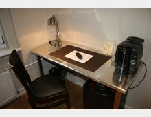 Silberner Schreibtisch mit einer schwarzen Ledersitzfläche auf einem Holzstuhl, einer metallenen Schreibtischlampe, einer Kaffeemaschine, einem Blatt Papier mit Fernbedienung und einer kleinen Broschüre. Die Umgebung zeigt helle Wände und einen Boden mit Holzoptik.