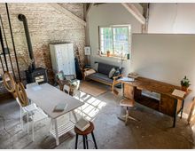 Offener Raum im rustikalen Stil mit einer Backsteinwand und Holzbalken. Zu sehen sind ein Holzofen, ein Holztisch mit Stühlen, ein Schreibtisch mit einem Hocker, ein Sofa vor einem großen Fenster sowie dekorative Gegenstände und Pflanzen.