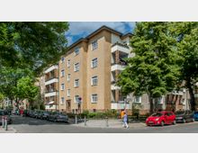 Wohnhaus mit vier Etagen und hellbrauner Fassade, ergänzt durch Balkone mit weißen Geländern und braunen Backstein-Elementen. Das Gebäude steht an einer von Bäumen gesäumten Straße mit parkenden Autos und einem Gehweg, auf dem Fußgänger unterwegs sind.
