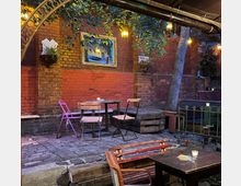 Ein kleiner, gemütlicher Außenbereich mit Backsteinwänden, vereinzelt aufgestellten Tischen und Stühlen, umgeben von Pflanzen und Beleuchtung durch hängende Laternen. Im Hintergrund sind ein Baum, ein dekorativer Rahmen an der Wand und hölzerne Elemente zu sehen.