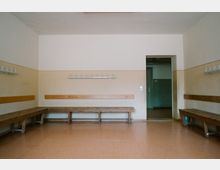 Ein schlichter Umkleideraum mit Holzbänken entlang der Wände, darauf befestigte Holzleisten und Haken zum Aufhängen. Der Raum hat helle Wände mit einem beigefarbenen Streifen und eine offene Tür führt in einen angrenzenden Bereich.
