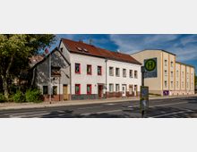 Wohnhäuser mit roten Fensterrahmen und Ziegeldächern an einer Straße; im Vordergrund steht ein Schild einer Bushaltestelle mit Fahrplan. Rechts ist ein modernes, gelb gestrichenes Gebäude zu sehen, links säumen Bäume den Gehweg.