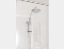 Eine verchromte Duscharmatur mit einer großen runden Regendusche und einer kleineren Handbrause, montiert an einer glatten weißen Duschwand. Im Hintergrund ist ein gerahmtes Bild undeutlich erkennbar.