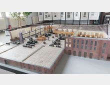 Modell eines historischen Fabrikgebäudes mit Backsteinfassade, offenen Innenräumen und detaillierter Nachbildung von Maschinen. Im Hintergrund sind ausgestellte Fotografien und Infotafeln in einem Ausstellungsraum sichtbar.