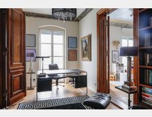 Arbeitszimmer mit einem großen Schreibtisch aus Metall und Glas, einem schwarzen Bürostuhl und Holzboden. Links und rechts stehen Lampen, an der Wand hängen mehrere Kunstwerke, und durch eine geöffnete Holztür ist ein weiterer Raum mit Kinderspielzeug sichtbar.