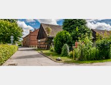 Eine kleine ländliche Straße mit alten Fachwerkhäusern und einem roten Backsteingebäude. Umgeben von Büschen, Bäumen und gepflegten Gärten mit blühenden Sträuchern, führt die Straße durch eine ruhige Dorfumgebung unter einem bewölkten Himmel.