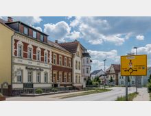 Straßenansicht in einer Kleinstadt mit mehrstöckigen Häusern im historischen Stil, darunter ein Gebäude mit dem Schriftzug „Bildungskooperation Thiele“. Im Vordergrund ein gelbes Verkehrsschild mit Wegweisern zu umliegenden Orten und einem Kreisverkehr.