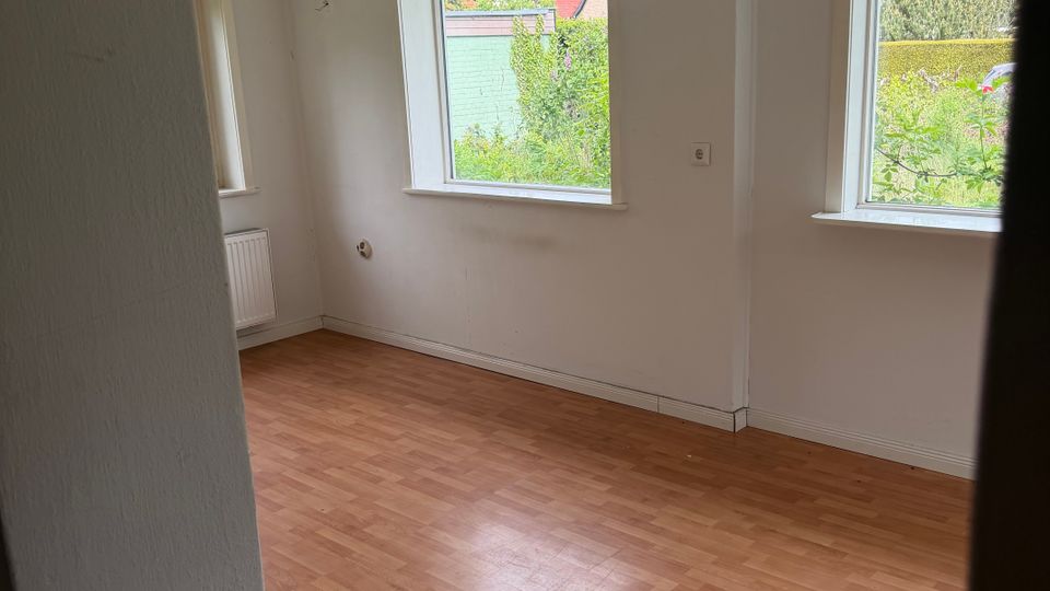 Leerer Innenraum mit hellem Laminatboden, weißen Wänden und freiliegendem Holzbalken unter der Decke. Zwei große Fenster lassen Tageslicht herein und bieten Blick ins Grüne sowie auf Nachbarhäuser.