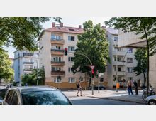 Straßenecke in einem Wohngebiet mit mehrstöckigen Wohnhäusern, großen Bäumen und Verkehrszeichen. Im Vordergrund stehen ein Fußgängerüberweg, geparkte Autos und eine Ampel.