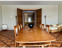 Esszimmer mit einem langen, hellbraunen Holztisch und passenden Stühlen mit gepolsterten Rückenlehnen. Im Hintergrund stehen geöffnete Doppeltüren, die in einen weiteren Raum mit Sofa, Sessel und Kronleuchter führen. Der Boden ist mit Holzdielen im Muster verlegt.