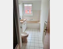 Kleines Badezimmer mit hellen Fliesen, Badewanne unter einem großen Fenster und Waschbecken mit Unterschrank auf der linken Seite. Im Vordergrund steht ein WC, rechts ist ein Heizkörper an der Wand, links ist die Kante einer Dusche erkennbar.