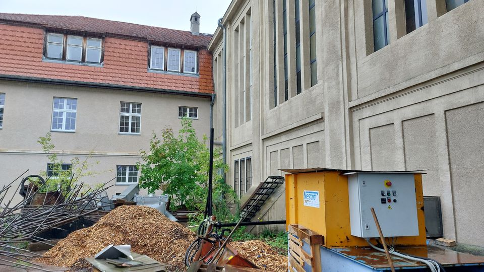 Außenansicht eines Gebäudekomplexes mit einer Fassade aus hellem Putz, großen vertikalen Fenstern und einem roten Ziegeldach. Im Vordergrund befinden sich Holzhackschnitzel, Metallstangen, ein gelber Maschinenkasten und etwas Grünbewuchs.