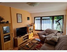 Wohnzimmer mit beigefarbenen Wänden, großen Fenstern mit weißen Vorhängen und Holzmöbeln, darunter ein TV-Schrank, eine Vitrine und ein Bücherregal. Im Raum stehen gepolsterte Sofas, ein Glastisch und ein Teleskop am Fenster.