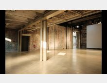 Industriehalle mit rohen Ziegelwänden, freiliegenden Metallträgern und einem glatten Betonboden. Im Raum befinden sich Metallpfeiler, eine geöffnete Tür im Hintergrund und eine weiße Projektionsfläche an der rechten Wand.