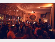 Ein Innenraum einer Bar oder eines Clubs mit dunkler, stimmungsvoller Beleuchtung und einer tanzenden Menschenmenge. An der Decke hängt eine Disco-Kugel, die Lichtpunkte an die Wände und Fenster wirft, während an der Rückwand eine Bar und Dekorationen sichtbar sind.