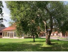 Ein weitläufiger Hof mit einem großen Baum im Vordergrund, umgeben von Gebäuden mit roten Ziegeldächern und verputzten Fassaden. Ein Teil der Gebäude hat große Fensterfronten, und der grüne Rasen dominiert die Szene.