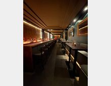 Schmaler, modern gestalteter Barbereich mit einer langen Theke auf der linken Seite und hohen Tischen mit Sitzbänken auf der rechten Seite. Die Decke und Beleuchtung sind dezent gestaltet, und Spiegel sowie indirektes Licht sorgen für eine warme Atmosphäre.