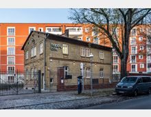 Zweistöckiges Gebäude aus gelbem Backstein mit Schriftzug „happy bite“ an der Fassade, umgeben von einem Zaun und einer gepflasterten Straße im Vordergrund. Im Hintergrund sind moderne, orangefarbene Wohngebäude und ein großer Baum sichtbar.