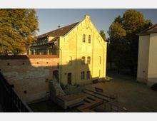 Gelbes Backsteingebäude mit Giebeldach und großen rechteckigen Fenstern, umgeben von Kopfsteinpflaster. Im Vordergrund eine historische Ziegelmauer mit Treppen und einem kleinen Wassergraben, im Hintergrund Bäume und weiteres Gebäude.