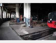 Innenansicht eines historischen Lokschuppens mit abgenutztem Betonboden und einem Wartungsgraben. Zahlreiche Arbeitsgeräte, Materialien und rote sowie schwarze Loks sind um eine zentrale Säule verteilt, während von der linken Seite Tageslicht durch hohe Fenster einfällt.