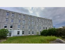 Ein mehrstöckiges Gebäude mit einer grauen Fassade aus Betonplatten und regelmäßigen rechteckigen Fenstern. Im Vordergrund befindet sich eine Grünfläche mit hohem Gras und vereinzelten Büschen, der Himmel ist bewölkt.