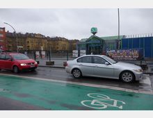 Straßenszene vor dem S-Bahnhof Hermannstraße in Berlin, mit einer Glasüberdachung am Eingang und einem grünen S-Bahn-Schild. Im Vordergrund sind parkende Autos und ein grüner Fahrradweg, im Hintergrund gelbe Wohngebäude und Werbeplakate sichtbar.