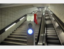 Treppenaufgang und Rolltreppe in einer U-Bahn-Station mit gefliesten Wänden und beleuchteten Handläufen. Im Vordergrund befindet sich eine Säule mit einem Leuchtknopf und einem roten Sicherheitskasten.
