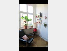 Ecke eines Wohnzimmers mit großen Fenstern, die Tageslicht einlassen. Rechts ein Wandregal mit Pflanzen und Dekoration, links ein schwarzer Ledersessel mit Kissen, daneben ein Tisch mit einer roten Tischlampe; eine große grüne Pflanze steht auf der Fensterbank.