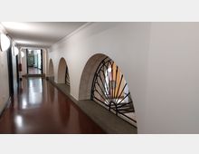 Ein heller Korridor mit glänzendem, rotbraunem Boden, weißen Wänden und gebogenen Metallgittern, die in Wandnischen eingelassen sind. Im Hintergrund ist ein angrenzender Raum mit einem Stuhl erkennbar.