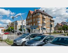 Straßenkreuzung in einer Stadt mit einem mehrstöckigen, historischen Gebäude mit gelblicher Fassade und roten Dachziegeln. Verkehrsschilder, parkende Autos und eine Ampelanlage prägen die Szene; im Hintergrund sind weitere ältere Gebäude und Bäume zu sehen.