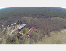 Luftaufnahme eines ländlichen Gebiets mit mehreren Gebäuden mit roten Dächern, umgeben von Wald und Wiesen. Im Hintergrund erstreckt sich ein dichter Nadelwald, während im Vordergrund vereinzelte Gebäude und Wege sichtbar sind.