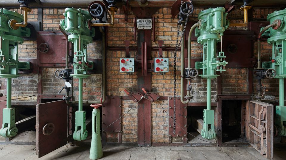 Industriehalle mit einer Ziegelwand, an der mehrere grüne, vertikale Maschinen mit Rohren, Ventilen und Schaltern befestigt sind. Unten sind offene Klappen und Metallstrukturen sichtbar, die zu einem alten mechanischen System gehören.