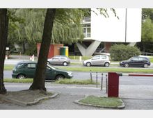 Ein moderner Gebäudekomplex mit auffallenden, schräg gestützten Säulen im unteren Bereich, umgeben von Grünflächen und Bäumen. Im Vordergrund sind parkende Autos entlang einer Straße sowie ein Gehweg mit Pollern und einer Schranke sichtbar.