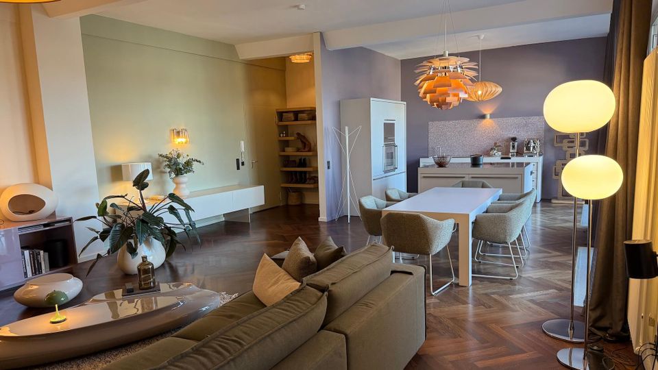 Open-plan modern living and dining room with a sofa and low coffee table leading to a long dining table and minimalist kitchen, all set on dark herringbone wood flooring. Pendant lights hang over the dining area, and large windows with curtains line one side of the room.