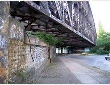Alte Eisenbahnbrücke mit rostiger Stahlkonstruktion und Graffiti, darunter ein gepflasterter Bürgersteig und eine Mauer aus Ziegelsteinen. Im Hintergrund befinden sich eine gepflasterte Straße, Bäume und geparkte Fahrzeuge.