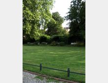 Grüne Parklandschaft mit einer großen, gepflegten Rasenfläche, umgeben von dichten Bäumen und Sträuchern. Im Hintergrund sind Blumenbeete und vereinzelte Parkbänke sichtbar.