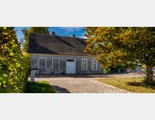 Ein kleines, graues Haus mit Satteldach und weißen Fenstern steht an einer gepflasterten Straße, umgeben von grünen Bäumen und Hecken unter einem klaren blauen Himmel.