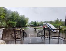 Eine Außenterrasse mit Metallgeländer und Treppe führt zu einem gepflasterten Hof, umgeben von Bäumen und Sträuchern. Im Hintergrund befinden sich ein kleines Gebäude, verschiedene Ausrüstungsgegenstände und eine Flugzeugtreppe, daneben steht ein roter Lkw.