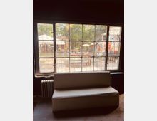 Ein großes Fenster mit Sprossen bietet den Blick auf einen Garten mit Sitzplätzen, Sonnenschirmen und bunten Wandgestaltungen im Hintergrund. Im Inneren des Raumes steht eine weiße Sofa-Bank vor dem Fenster, daneben ist ein Heizkörper sichtbar.